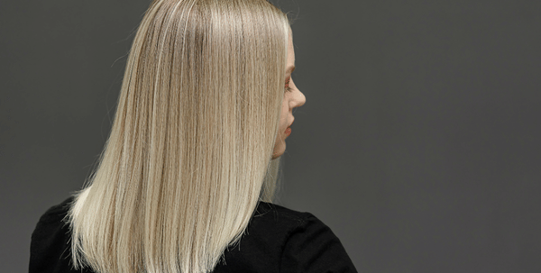 O femeie cu păr blond, drept, până la umeri, este surprinsă din spate; poartă un top negru și stă cu fața spre un fundal gri, simplu.