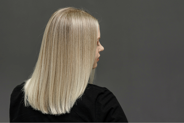 O femeie cu păr blond, drept, până la umeri, este surprinsă din spate; poartă un top negru și stă cu fața spre un fundal gri, simplu.